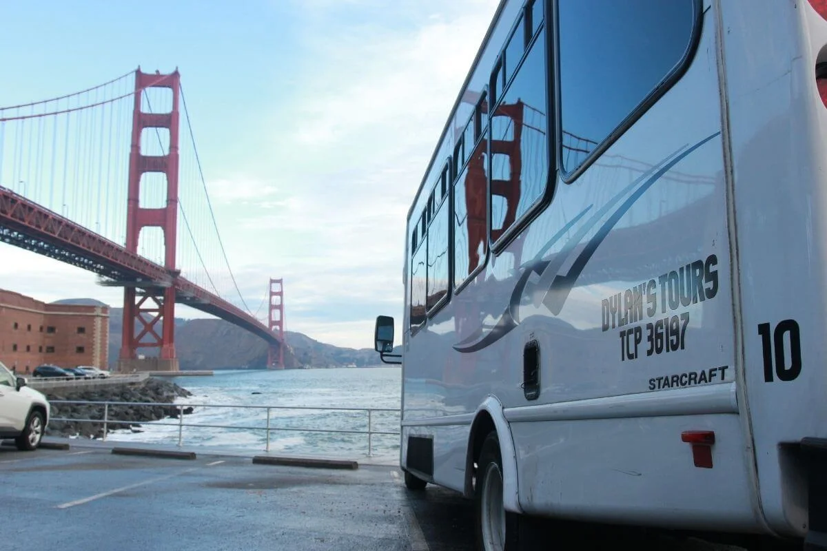 Tour bus parked near the Golden Gate Bridge in San Francisco with the bridge reflected in the bus windows and the bay in view