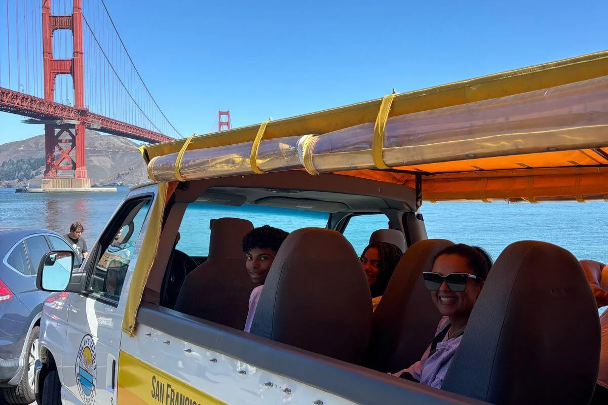 Passengers smiling inside an open-air tour vehicle parked near the Golden Gate Bridge on a sunny day in San Francisco.