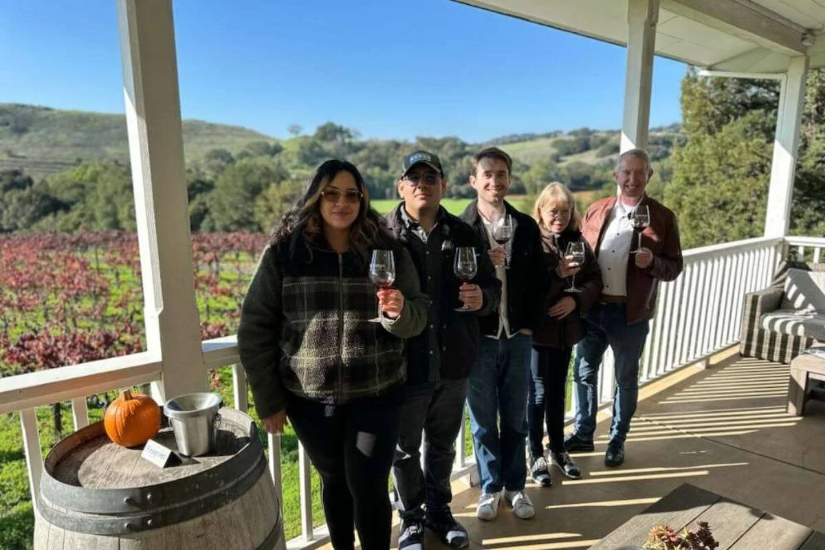A group of people holding wine glasses on a covered porch overlooking green vineyards and rolling hills on a sunny day.