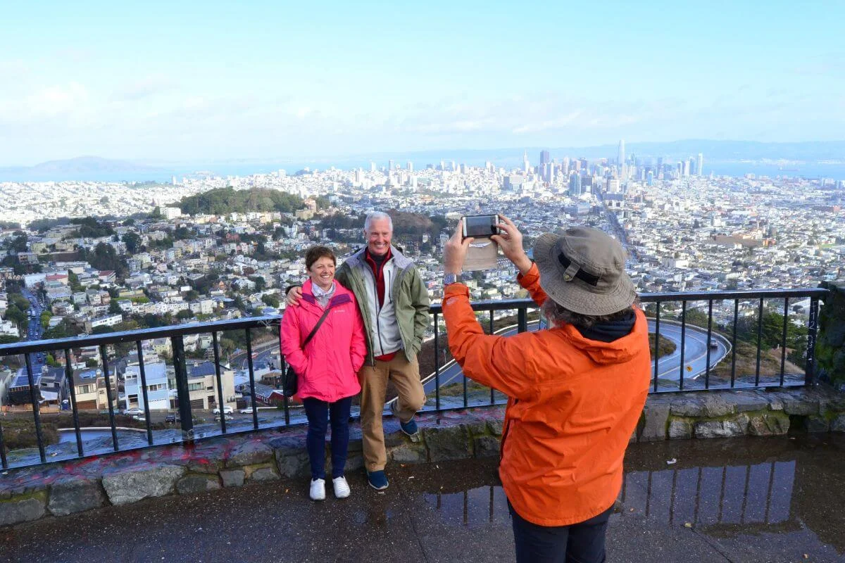 A couple poses for a photo at a scenic overlook while another person takes their picture, with the San Francisco skyline and city neighborhoods spread out below.
