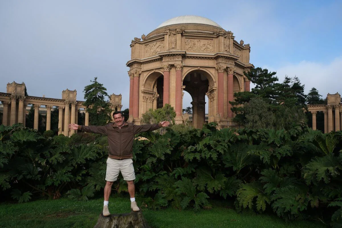 A man stands with his arms outstretched in front of the Palace of Fine Arts, surrounded by greenery and the historic domed structure in San Francisco.