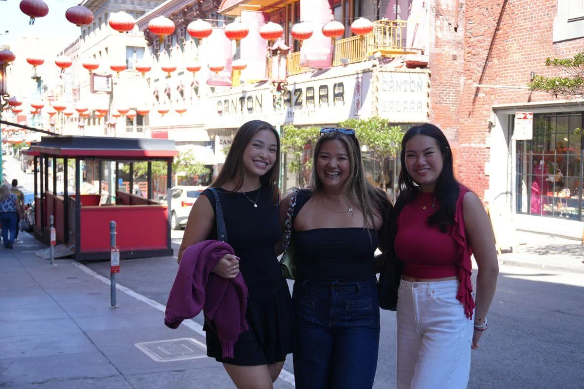 Three women smiling and posing together on a Chinatown street in San Francisco, with red lanterns hanging overhead, storefronts in the background, and a cable car stop nearby.