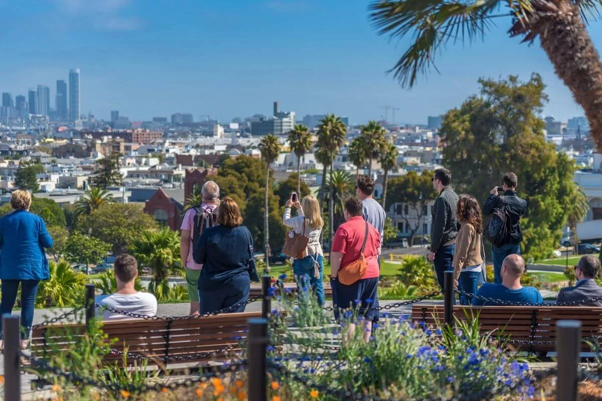People standing and sitting at Dolores Park overlooking the San Francisco skyline on a sunny day