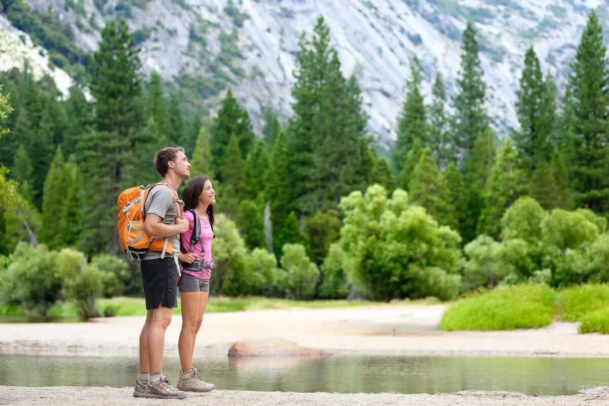 Two hikers standing by a river with backpacks surrounded by pine trees and mountains in Yosemite National Park
