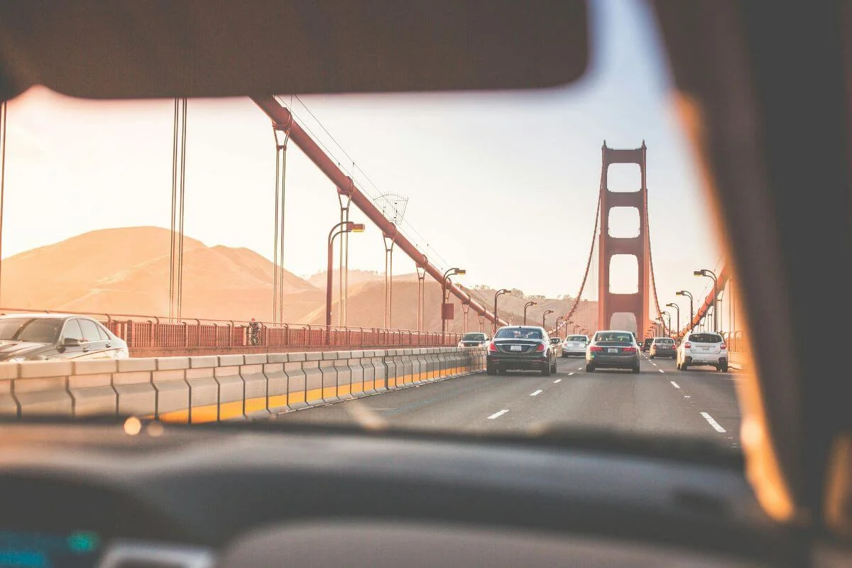 A view from inside a car driving across the Golden Gate Bridge in San Francisco, with vehicles ahead, the bridge’s red towers rising in the distance, and warm sunlight illuminating the surrounding hills.