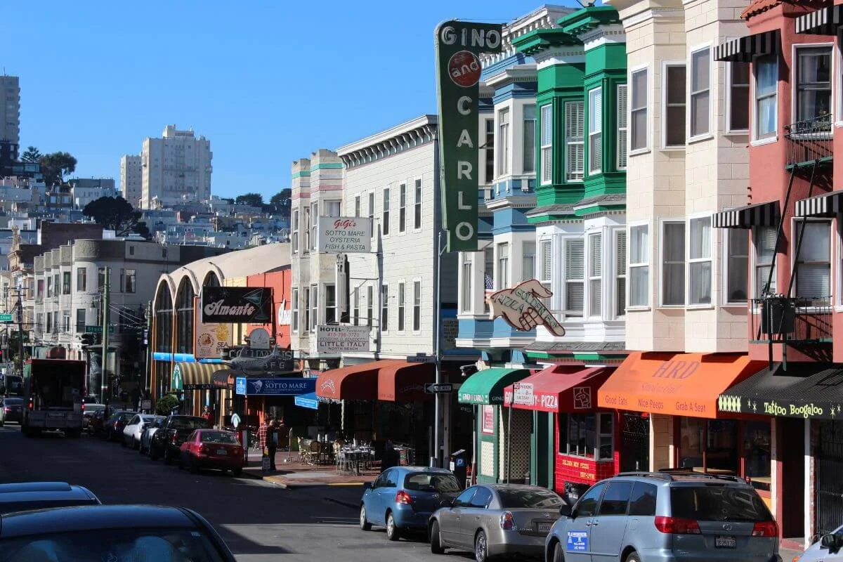 A colorful street in San Francisco’s Little Italy (North Beach), lined with Italian restaurants, cafés, and storefronts, with parked cars along the road and hillside buildings in the background under a clear blue sky.
