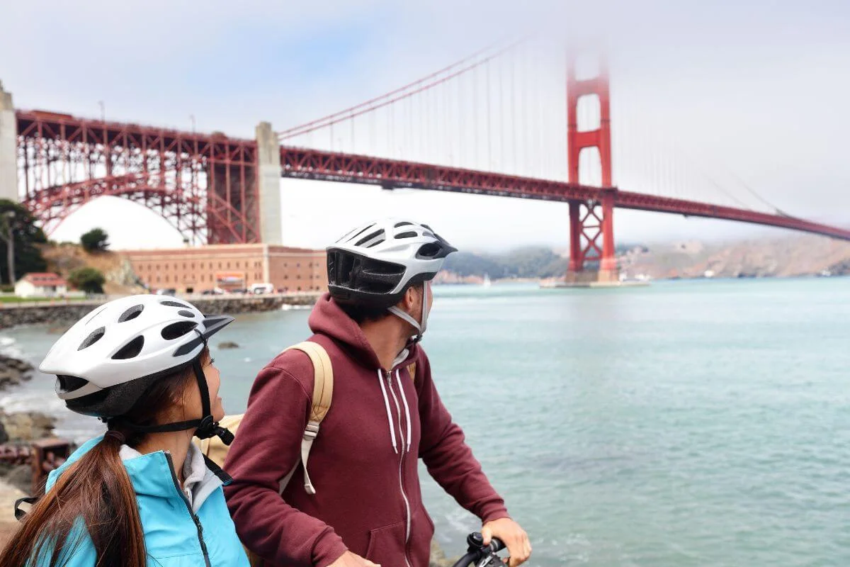Two cyclists wearing helmets stop along the waterfront to view the Golden Gate Bridge and San Francisco Bay on a clear day.