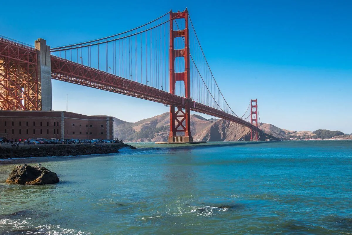 The Golden Gate Bridge spanning San Francisco Bay on a clear day, with blue water in the foreground and rolling hills in the background.