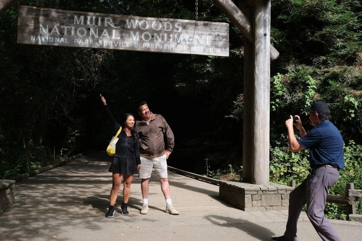 A couple poses beneath the Muir Woods National Monument entrance sign while another visitor takes their photo along the forest walkway.