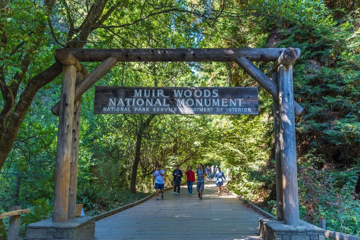 Wooden entrance sign for Muir Woods National Monument with visitors walking along a shaded boardwalk surrounded by lush redwood forest.