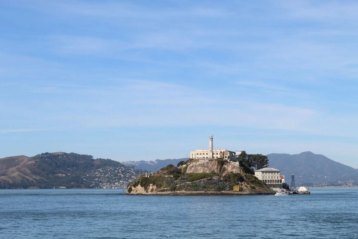 Alcatraz Island viewed from the water, showing the historic prison buildings and lighthouse against a calm San Francisco Bay with hills in the background.