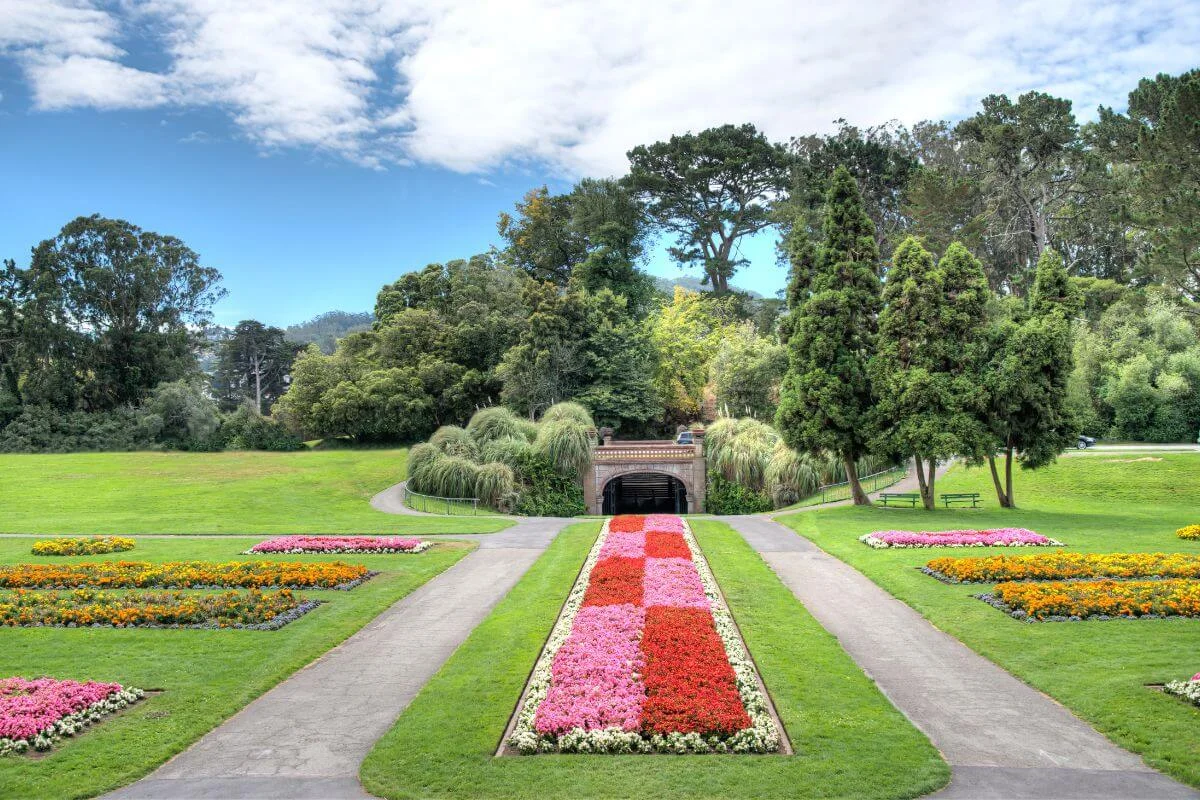 A symmetrical garden landscape with manicured lawns, bright flower beds, and a central floral walkway leading to a tunnel surrounded by trees.
