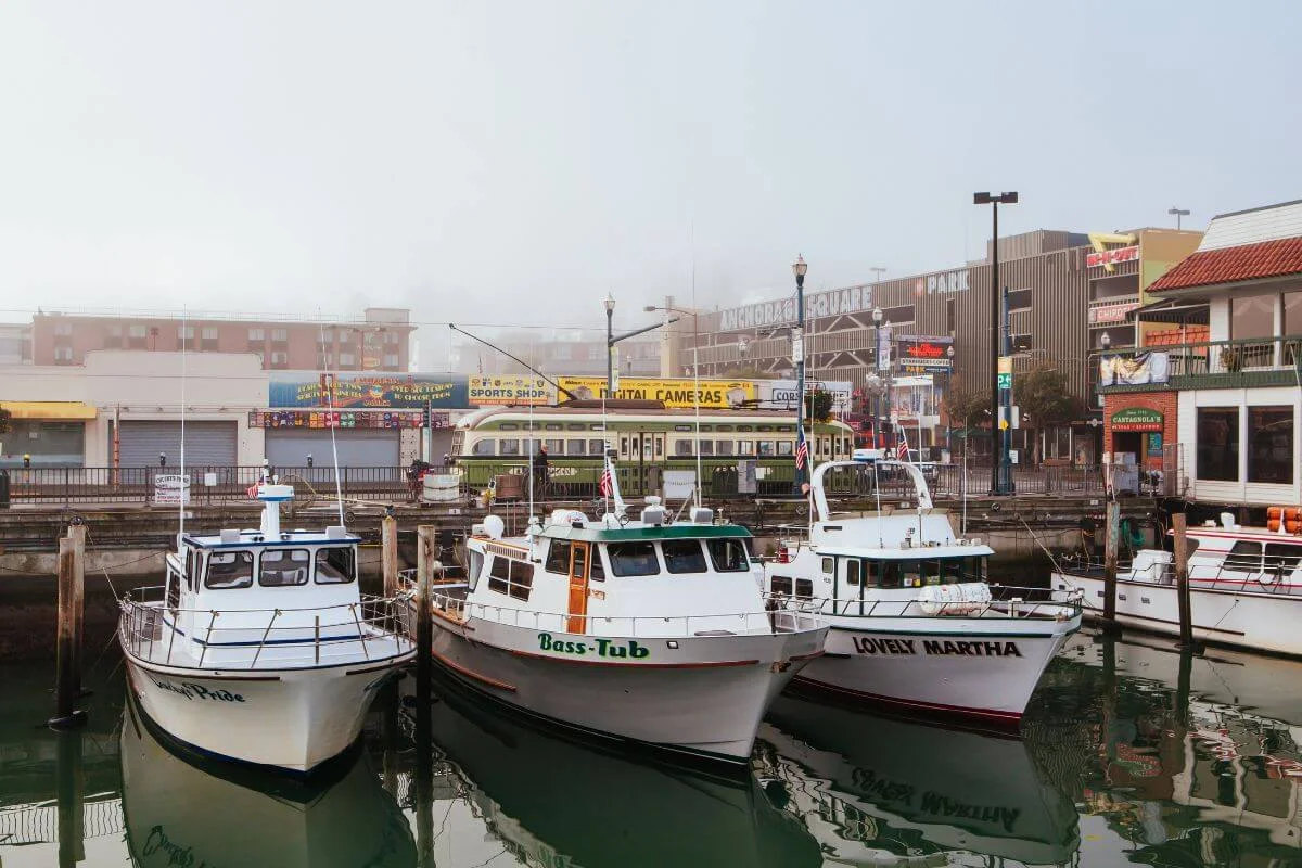 Fishing boats docked at Fisherman’s Wharf with storefronts, signage, and a historic streetcar visible in the background on a foggy San Francisco day.