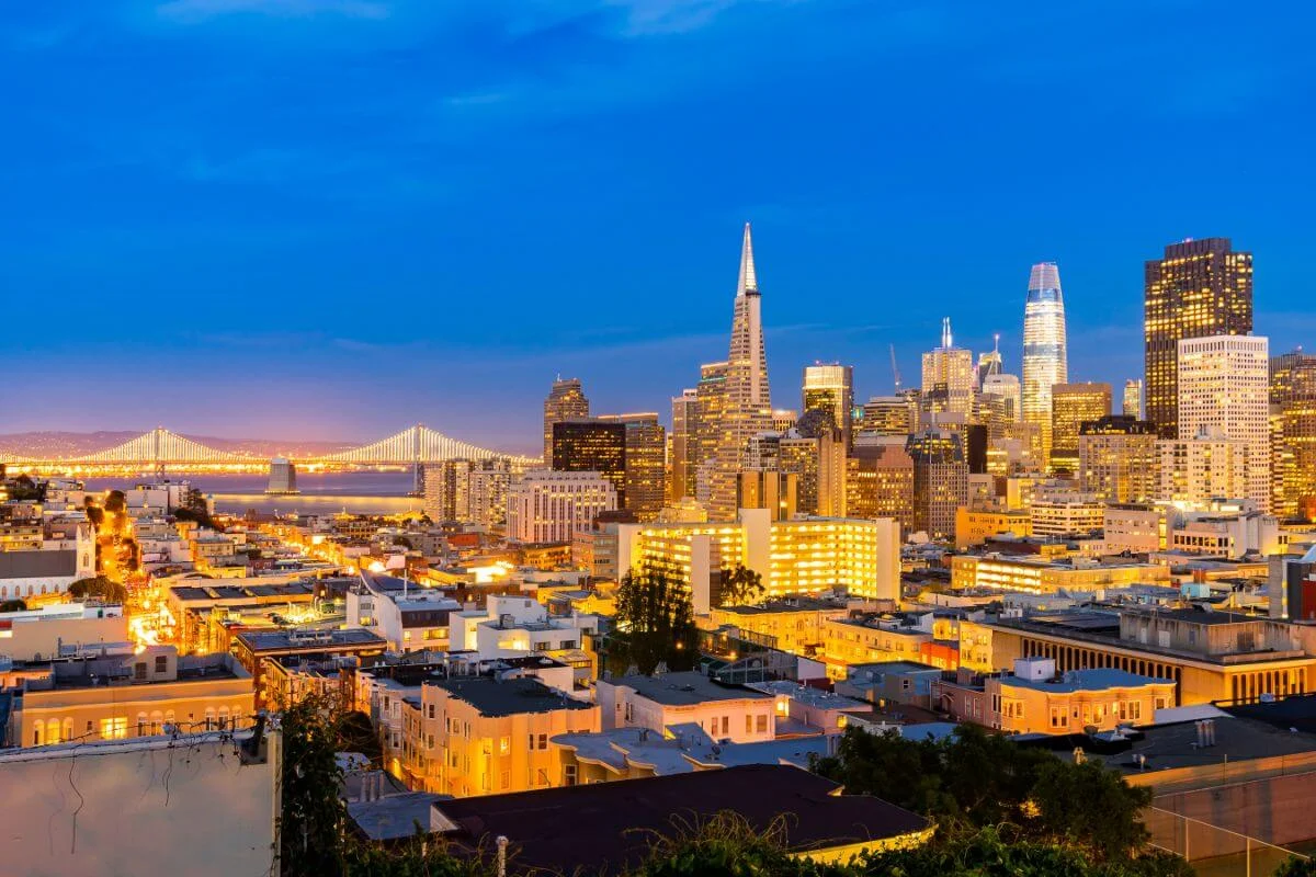 Evening view of the San Francisco skyline with illuminated skyscrapers, city lights, and the Bay Bridge visible against a deep blue sky