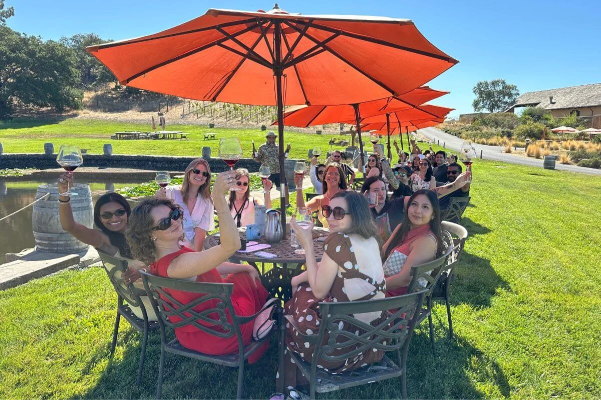 Group of visitors enjoying an outdoor wine tasting under orange umbrellas at a Napa Valley vineyard lawn seating area