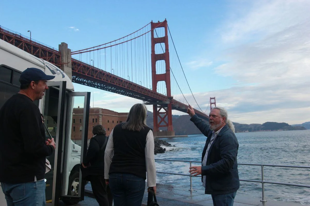 Tour guide pointing toward the Golden Gate Bridge while guests stand beside a tour van at the waterfront in San Francisco