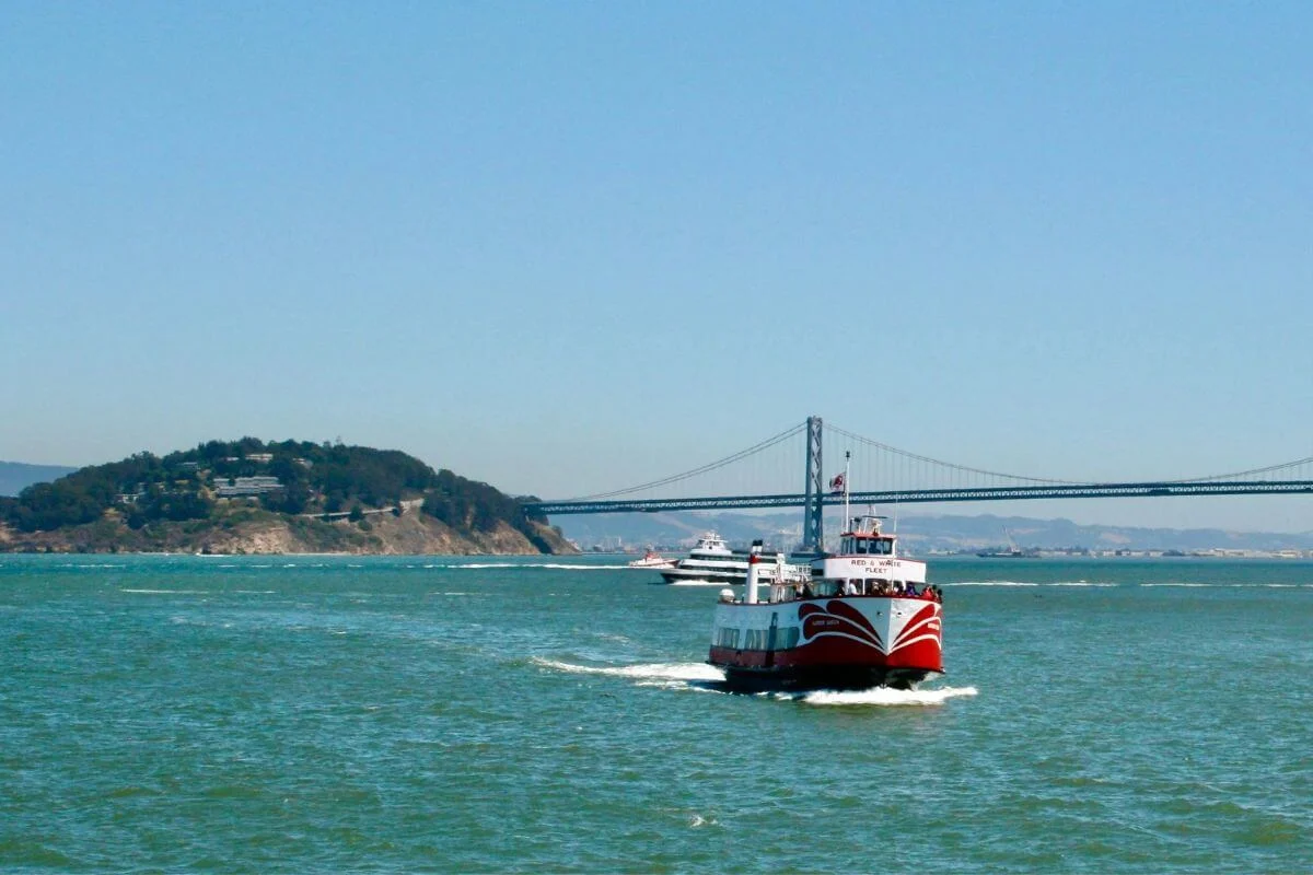Red and white ferry boat cruising across San Francisco Bay with the Bay Bridge and Yerba Buena Island in the background.