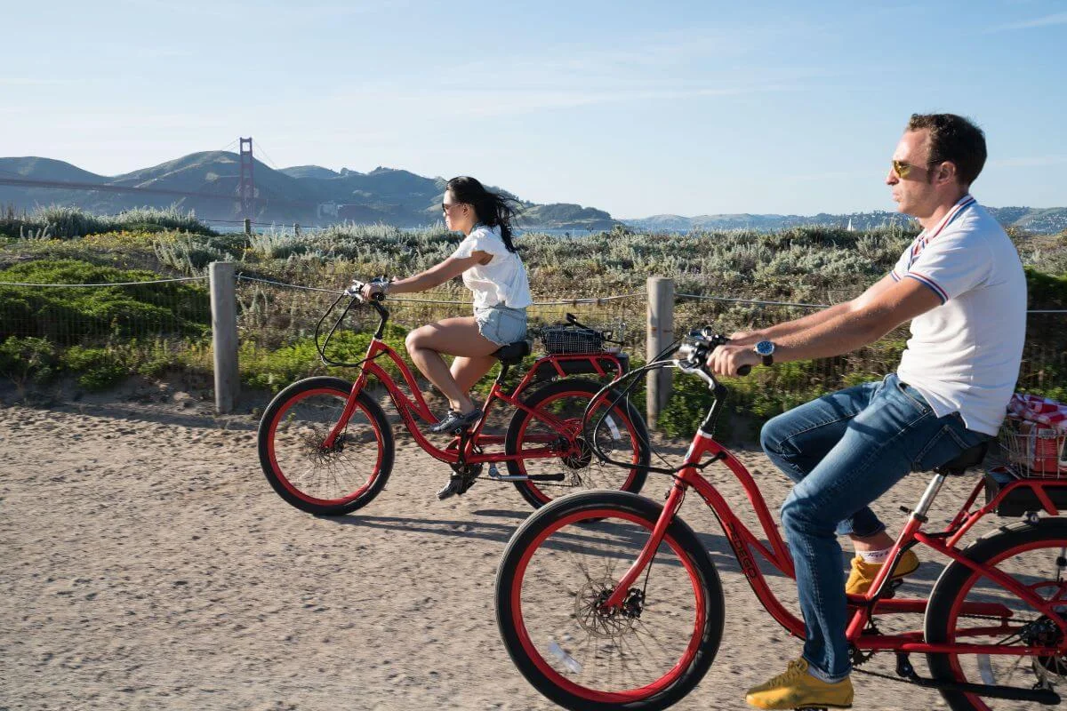 Two adults riding red bicycles along a coastal trail with the Golden Gate Bridge visible in the background in San Francisco.