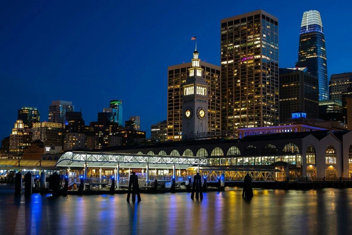 Night view of the San Francisco Ferry Building with its illuminated clock tower, surrounding downtown skyscrapers, and reflections on the bay along the Embarcadero