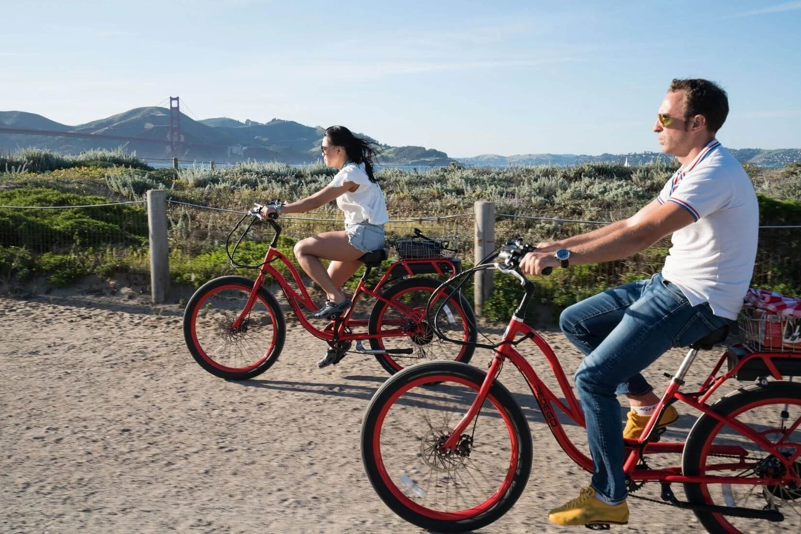 Two people riding red bicycles along a coastal trail in San Francisco with the Golden Gate Bridge and rolling hills visible in the background on a clear day