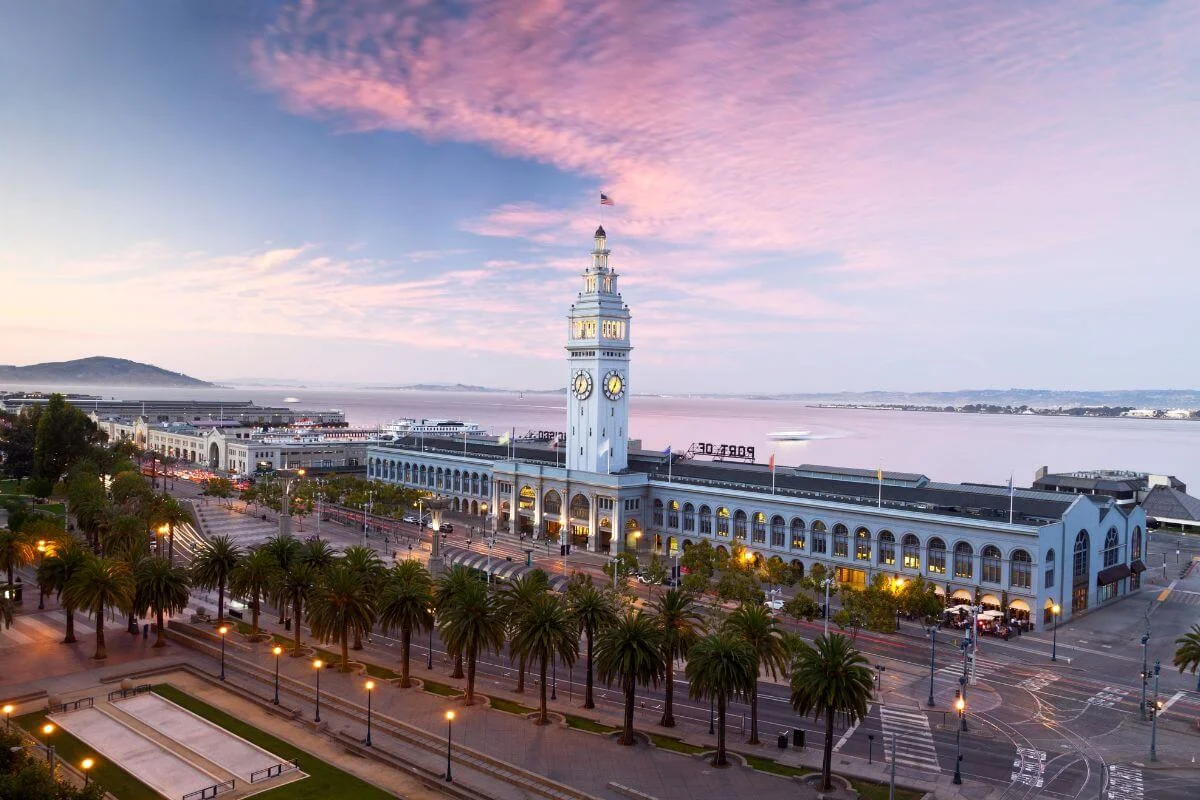 Aerial view of the Ferry Building in San Francisco with its iconic clock tower along the Embarcadero waterfront at sunset, palm trees lining the street and the bay in the background