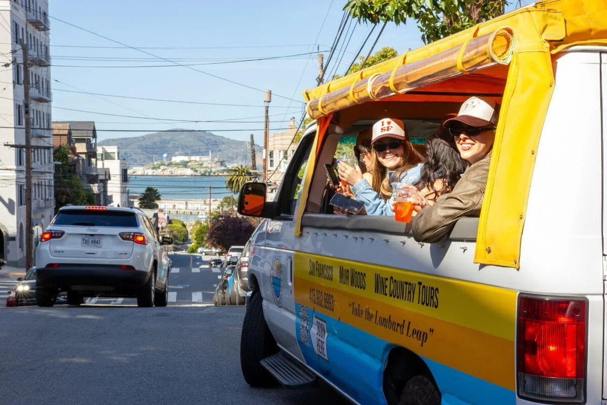 Passengers riding in an open air tour vehicle along Lombard Street in San Francisco with city hills, nearby buildings, and the bay visible in the background on a sunny day