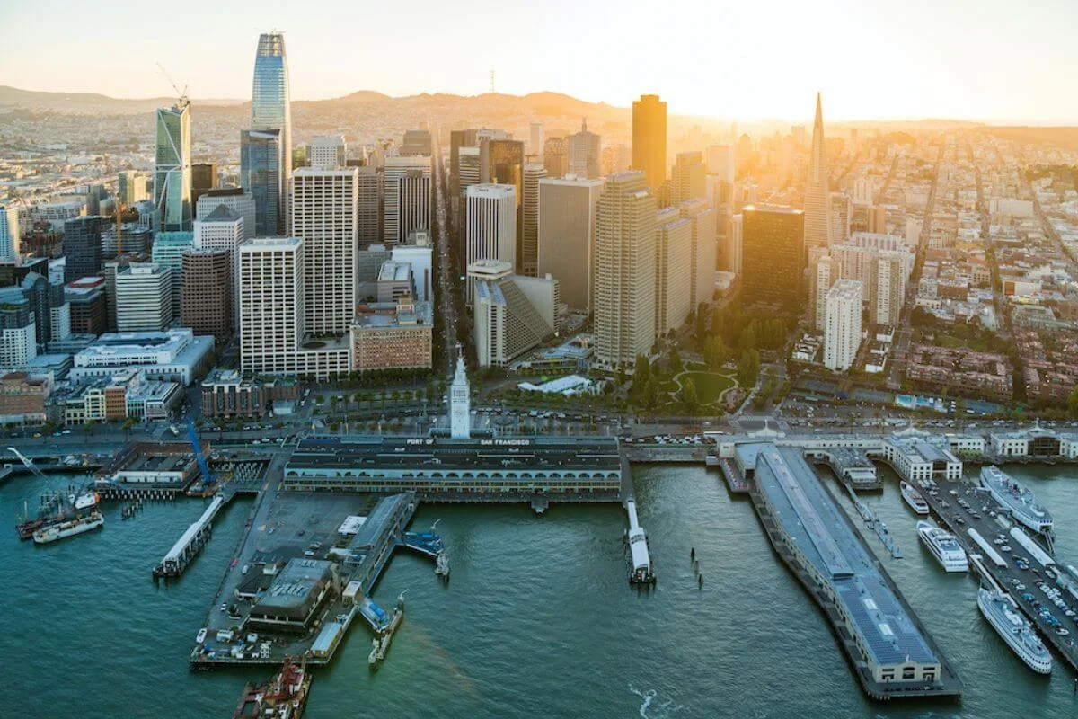 Aerial view of the San Francisco Ferry Building along the Embarcadero with downtown skyscrapers, piers, and the bay visible during golden hour