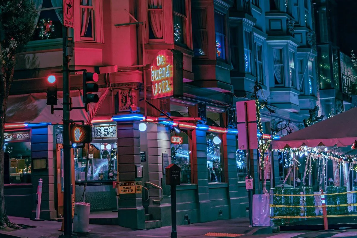 A colorful neon-lit restaurant on a city street corner at night, with bright red and blue lights, holiday decorations, and a traffic signal illuminating the urban scene.