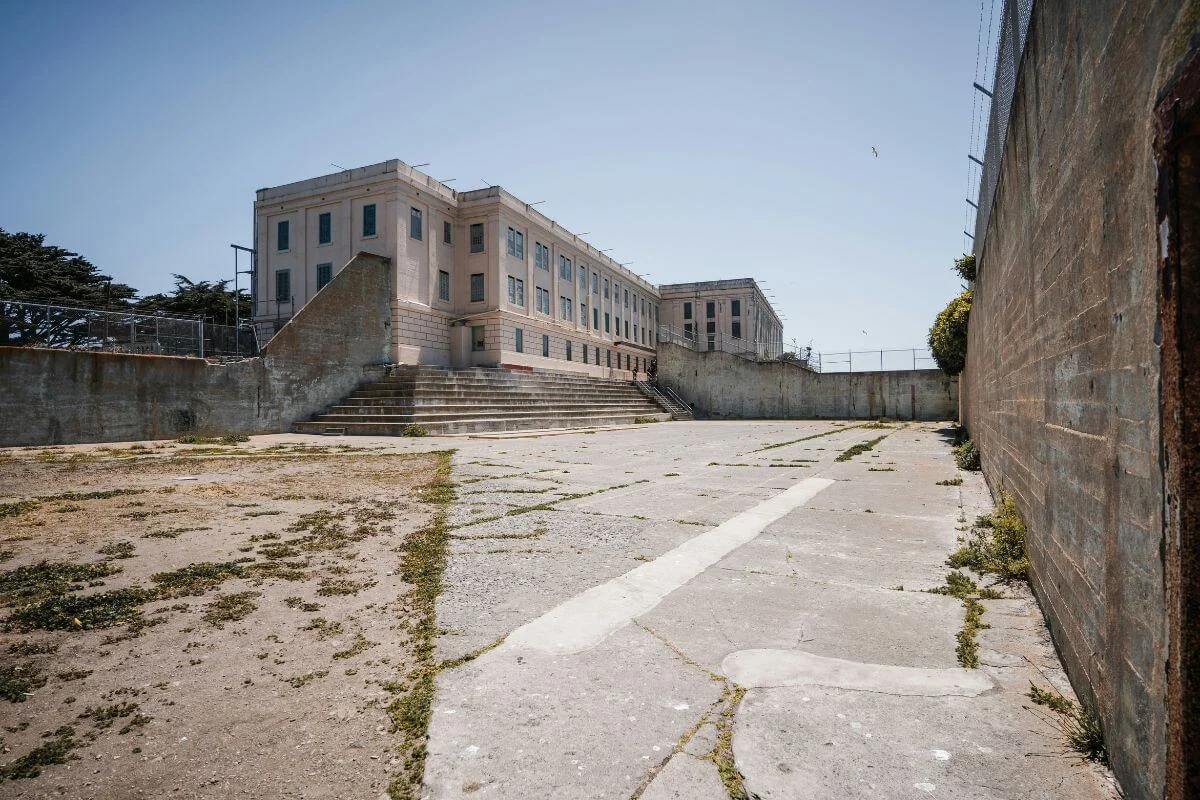 An open concrete prison yard on Alcatraz Island leading toward the main cellhouse building, surrounded by high walls and fences under a clear sky.