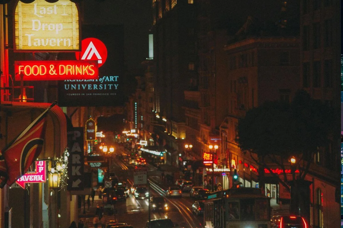 A busy city street at night filled with glowing neon signs, restaurants, cars, and streetlights creating a vibrant nightlife atmosphere.