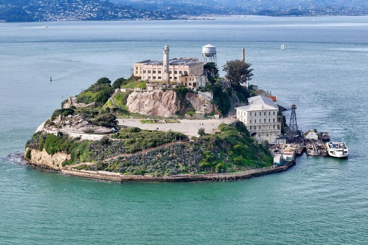 An aerial view of Alcatraz Island showing the historic prison buildings, lighthouse, dock, and surrounding blue waters with the San Francisco shoreline in the distance.