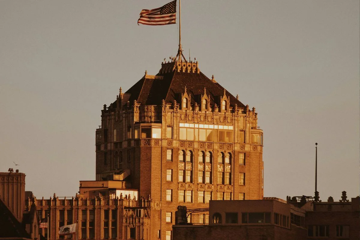 A historic multi-story building with ornate architecture and an American flag on top, illuminated by warm golden sunset light against a clear sky.