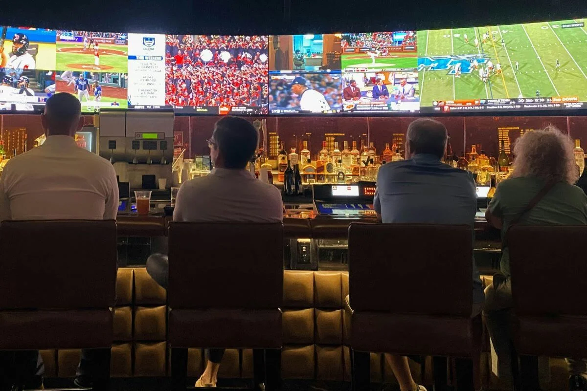 People seated at a bar watching multiple sports games on large screens while drinks and bottles line the counter in a lively sports bar atmosphere.