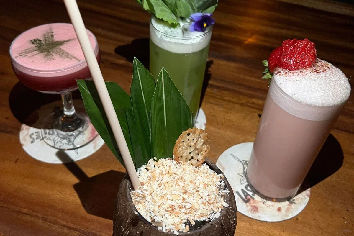A selection of colorful handcrafted cocktails on a wooden bar table, including a coconut drink topped with toasted coconut flakes, a strawberry milkshake-style cocktail, and vibrant garnished drinks with herbs and fruit.