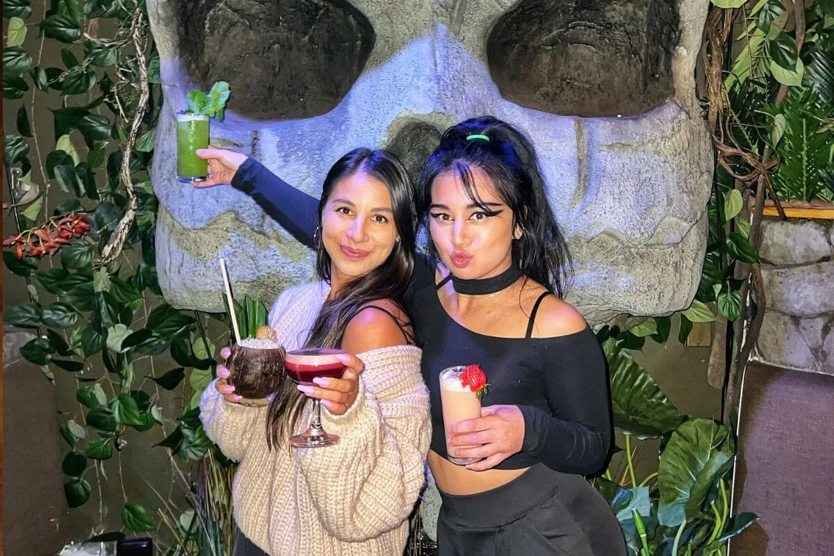 Two friends posing with colorful cocktails in front of a large stone sculpture surrounded by lush plants at a vibrant tropical-themed bar.