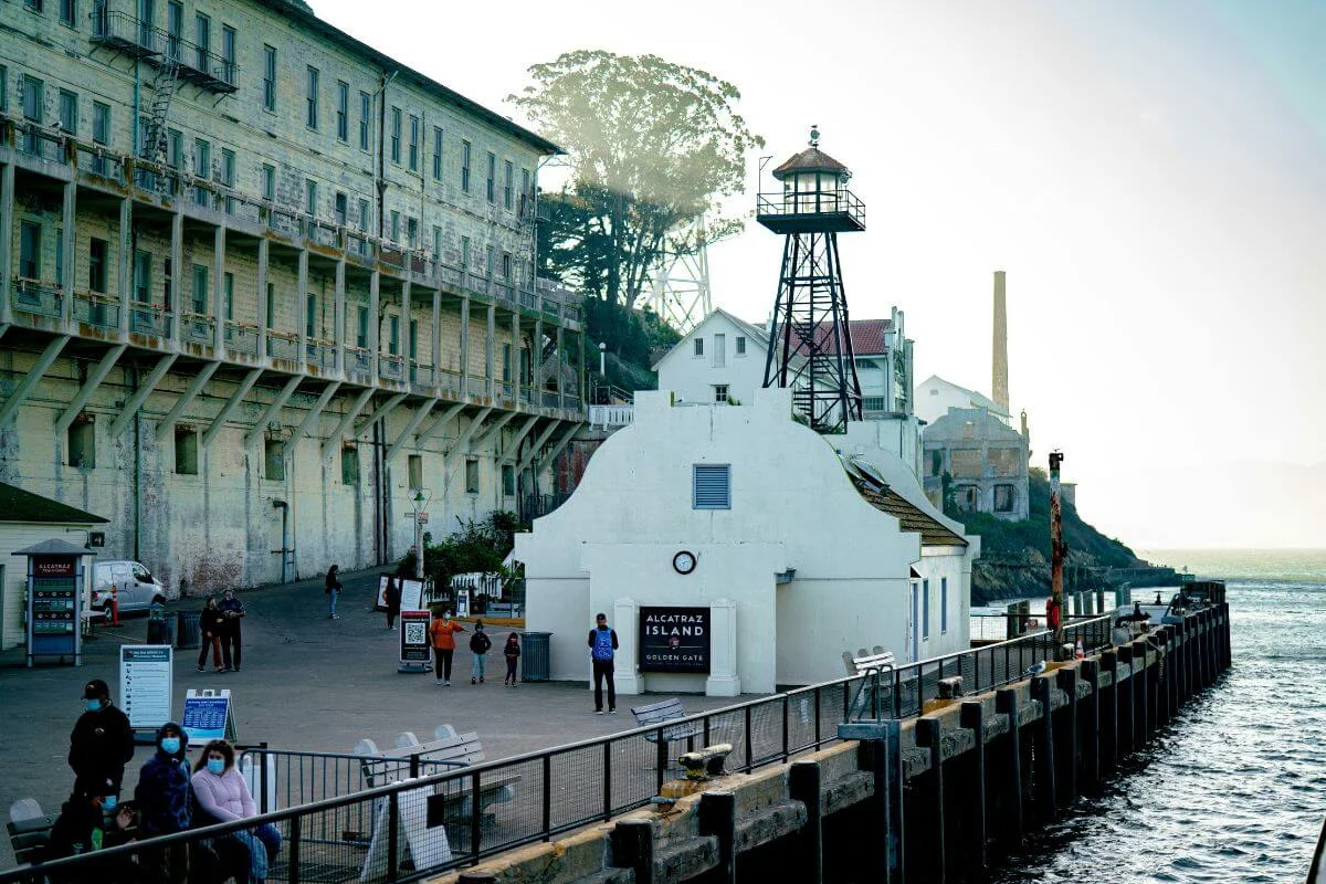 Visitors walking near the dock on Alcatraz Island with historic buildings and a guard watchtower overlooking the water along the San Francisco Bay.