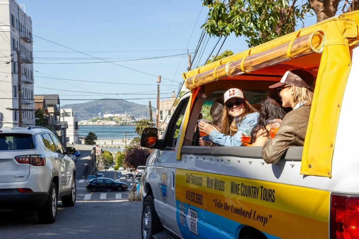 Tourists enjoying a ride in an open-sided tour van driving through San Francisco streets with views of the bay and city in the background.