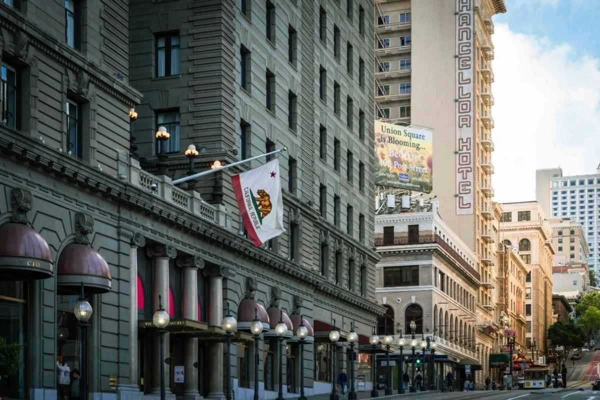 Street scene near Union Square in San Francisco featuring historic buildings, hotel facades, California flag, and a cable car in the distance.