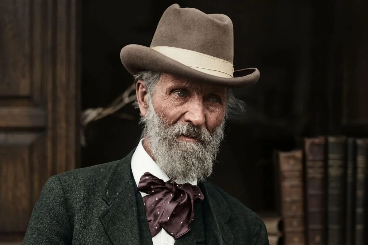 Close-up portrait of John Muir wearing a hat and bow tie, showcasing his iconic beard and thoughtful expression in a historical setting.