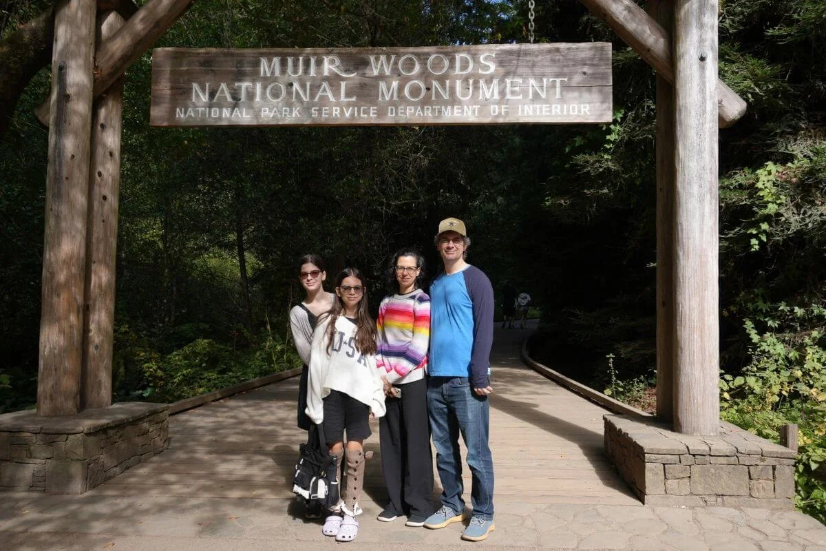 Family posing under the Muir Woods National Monument wooden entrance sign surrounded by towering redwood forest in California.