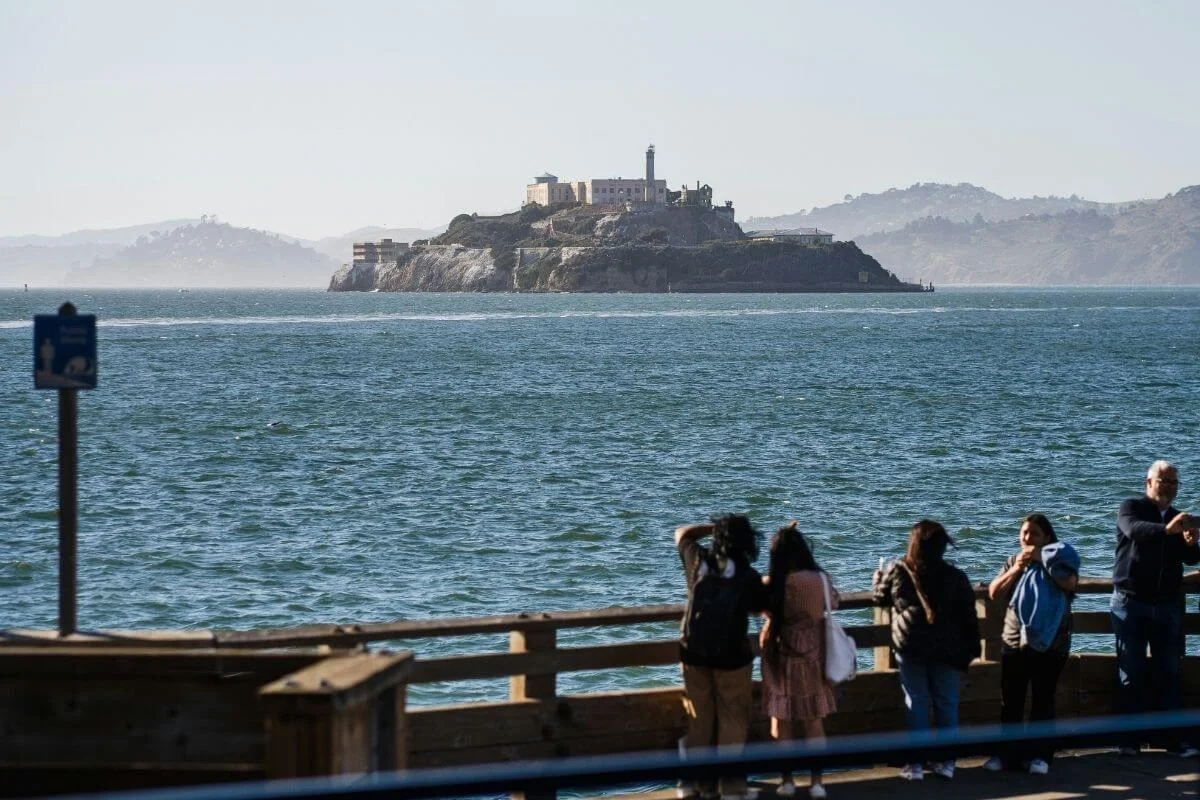 People standing along a waterfront pier in San Francisco looking out at Alcatraz Island across the bay on a clear day.