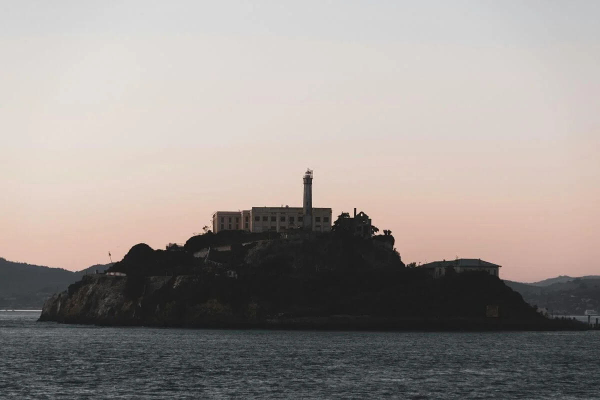 Moody silhouette of Alcatraz Island rising from the dark waters of San Francisco Bay at sunset, with the historic prison building and lighthouse visible against a soft pink sky.