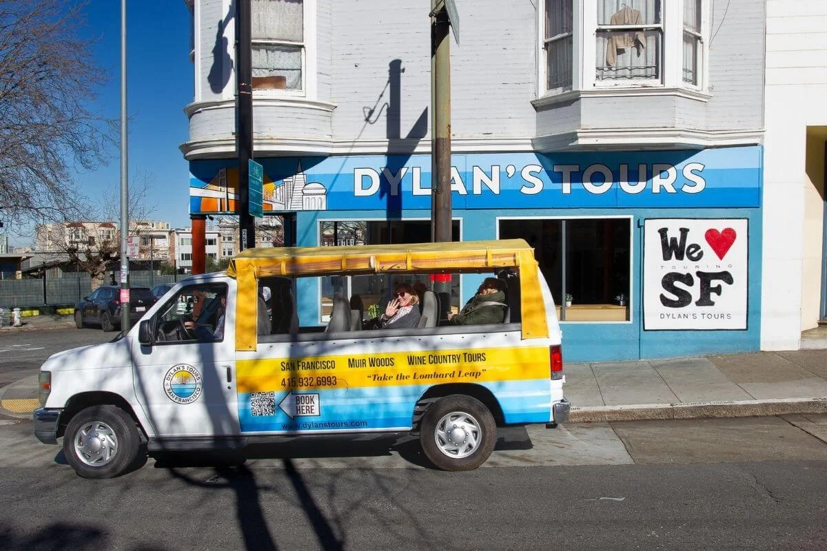 Small tour van parked outside Dylan’s Tours office in San Francisco with passengers inside, promoting city, Muir Woods, and wine country tours.