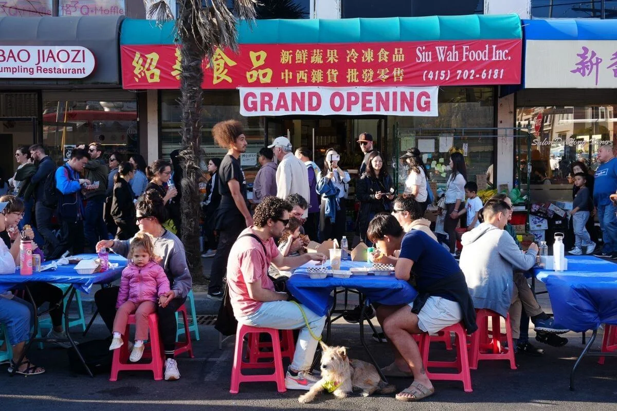People enjoying outdoor dining at a lively Chinatown street market in San Francisco, with local vendors, colorful signage, and visitors gathered around tables.