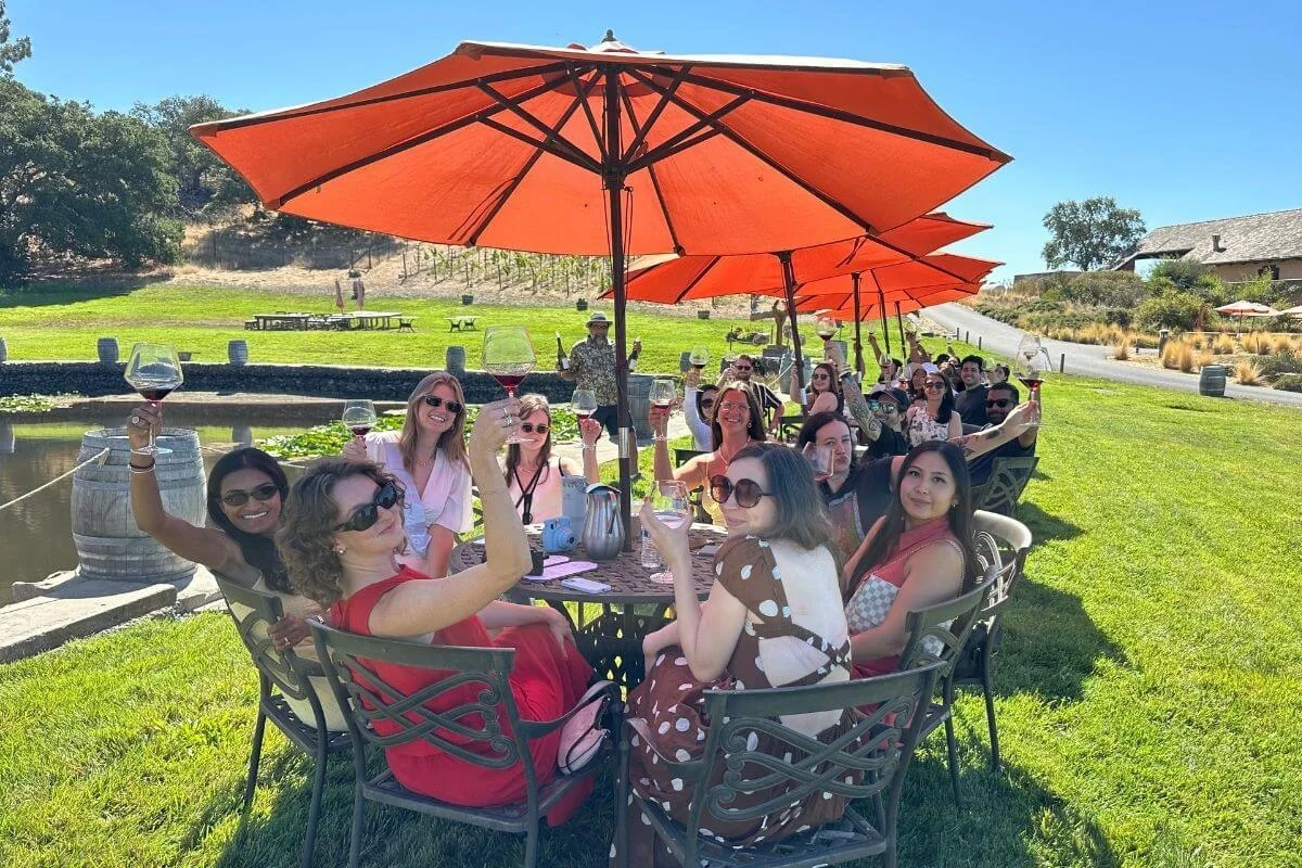 A large group of smiling visitors raising their wine glasses in a toast while seated at outdoor tables under orange umbrellas at a Napa Valley winery