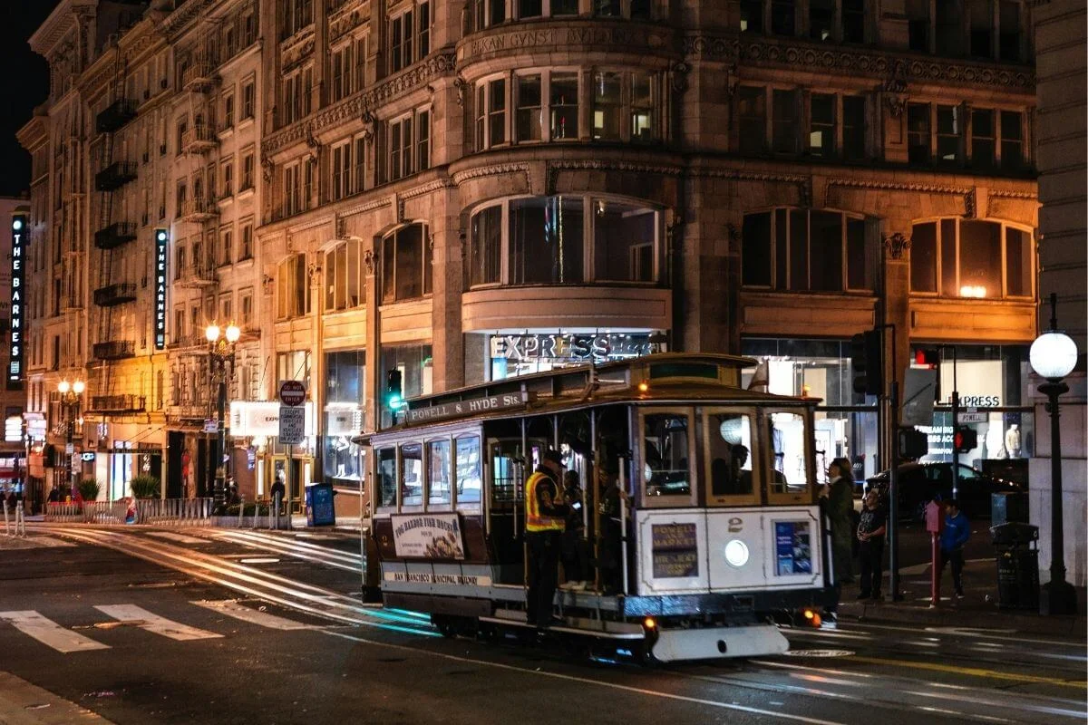 Historic Powell and Hyde Street cable car navigating a busy downtown San Francisco intersection at night, surrounded by lit storefronts and ornate early 20th century buildings.