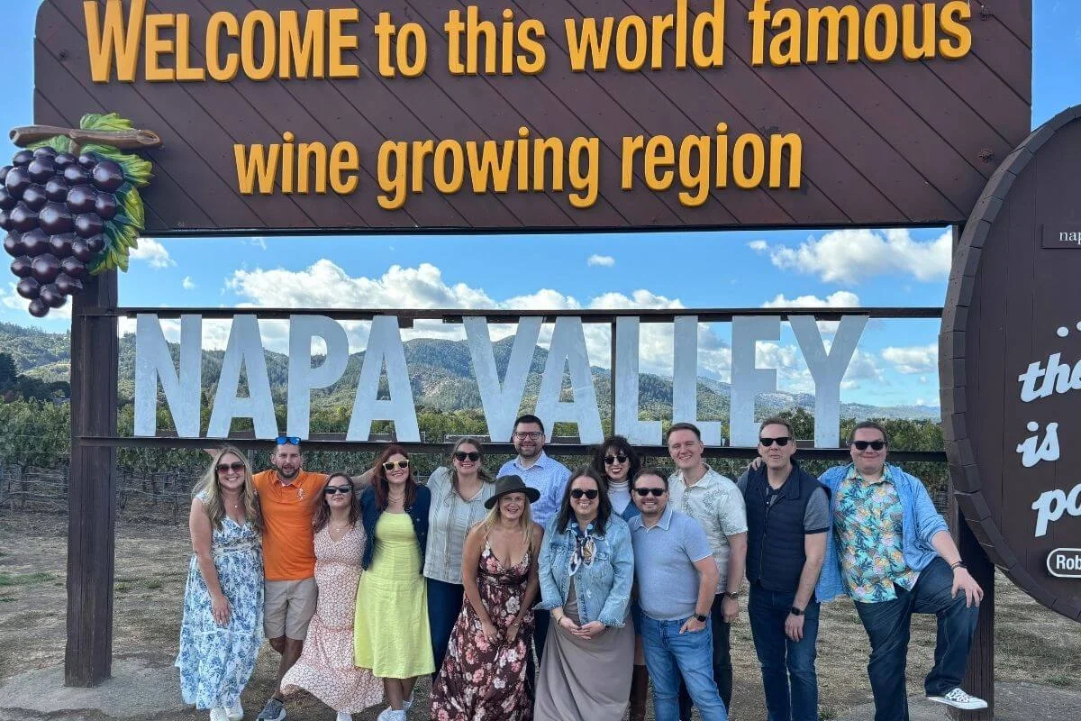 A group of twelve people smiling and posing together under the iconic Napa Valley welcome sign with vineyard-covered hills in the background