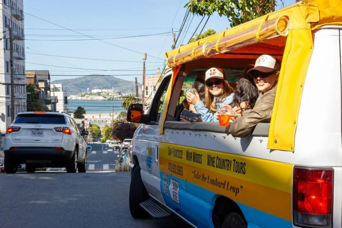 Tourists riding in a colorful tour van descending Lombard Street area in San Francisco with city views and waterfront in the distance.