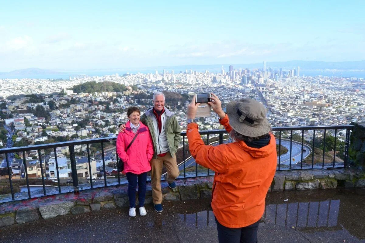 Tourists taking photos at Twin Peaks viewpoint with panoramic views of the San Francisco skyline and surrounding neighborhoods.