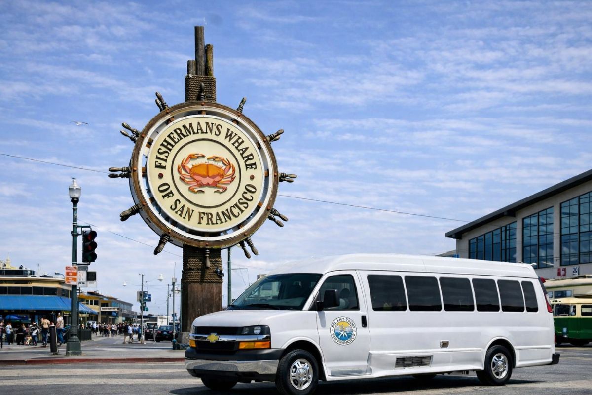 A vintage cream and red F Market streetcar headed to Fisherman's Wharf passes the iconic ship wheel landmark sign on a sunny day, with crowds of tourists gathered on the sidewalk and the Ripley's Believe It or Not building visible on the right.